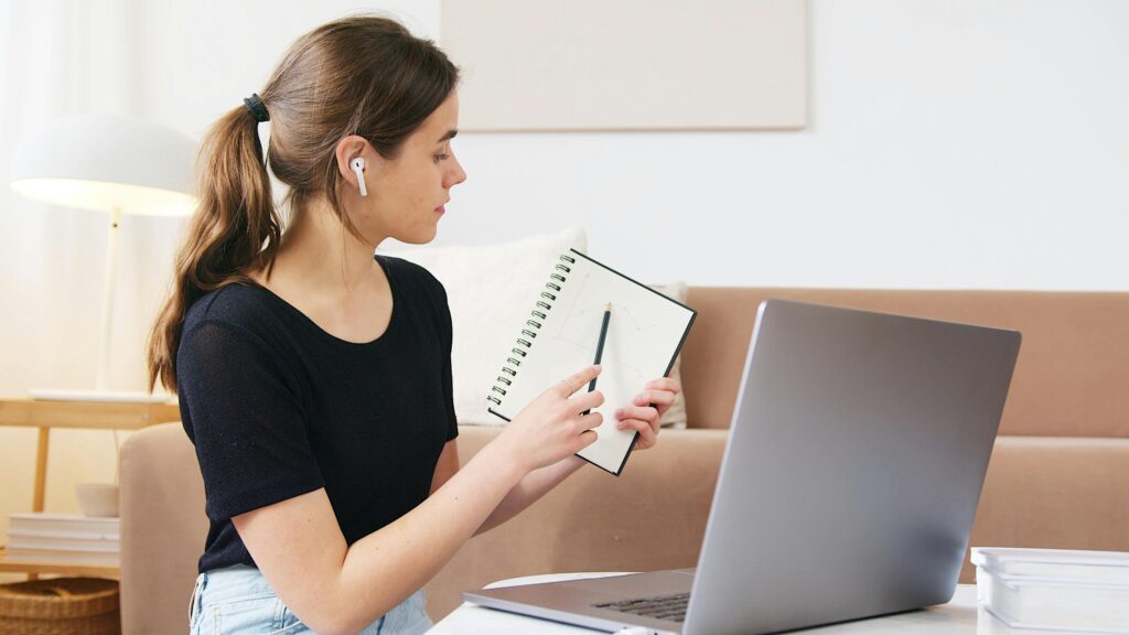 Concentrated young female student in earbuds showing notebook with notes during video conference distance learning concept