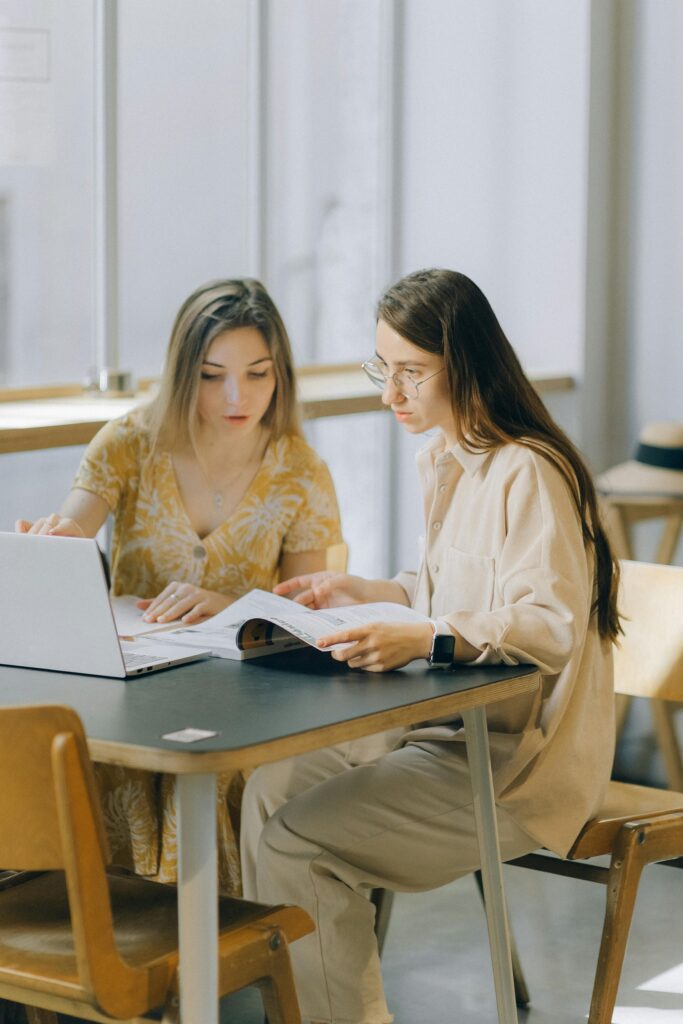 Two women engaged in study, using a laptop and book in a bright indoor setting.