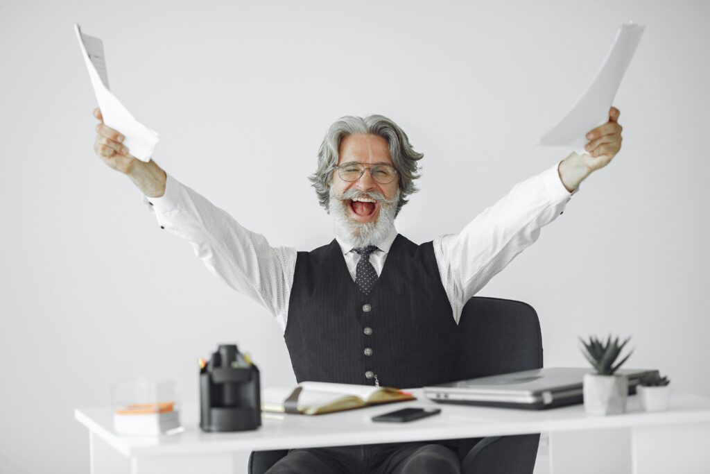 Joyful elderly man at desk celebrating success with raised arms holding papers, smiling confidently.