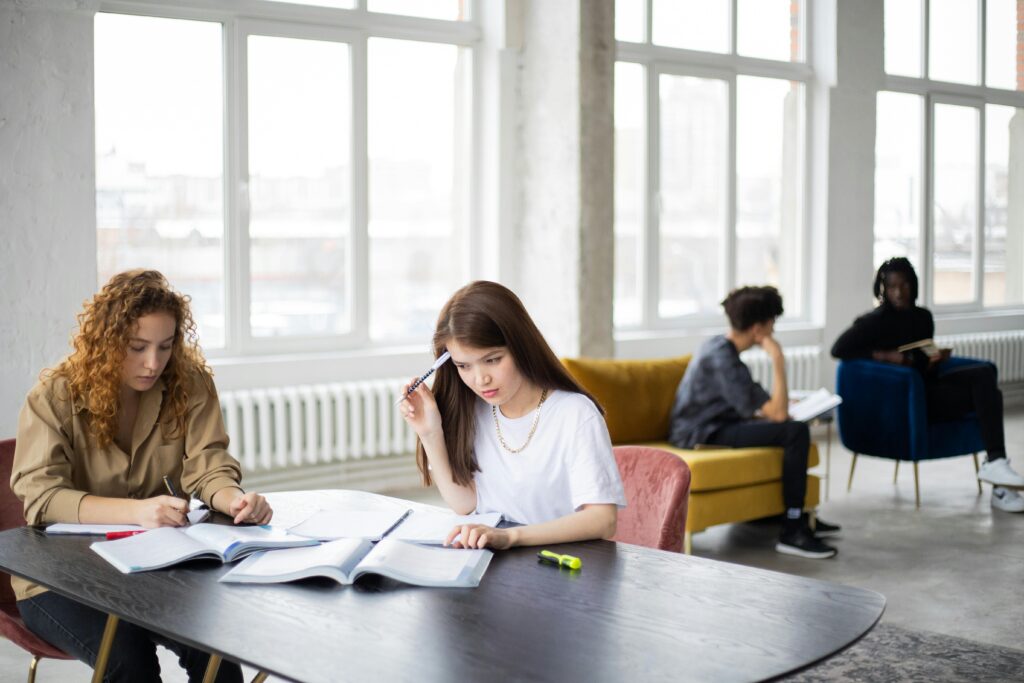 Pensive female reading book while sitting with friend and studying together against blurred guys communicating in modern workspace in daytime